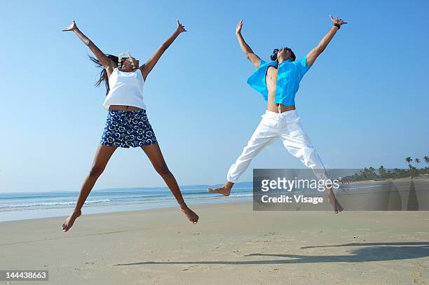 couple jumping on a beachside - mann freudensprung sonne vorderansicht leger stock-fotos und bilder