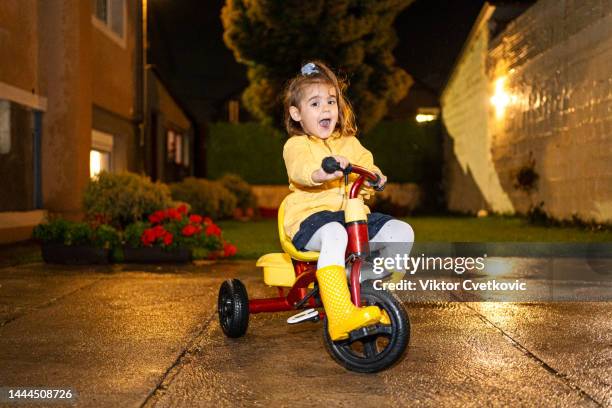 little girl dressed in yellow ride bicycle in the yard during the raining evening - girl rain night stock pictures, royalty-free photos & images