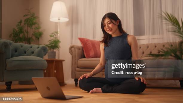 pregnant woman sitting on floor and stretching her body while watching online tutorials on laptop in living room at home - warm light stock pictures, royalty-free photos & images