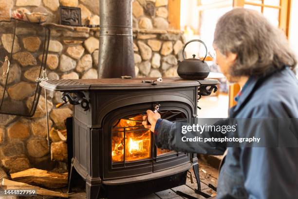 a senior man putting firewood his fireplace as winter arrives - wood burning stove stock pictures, royalty-free photos & images