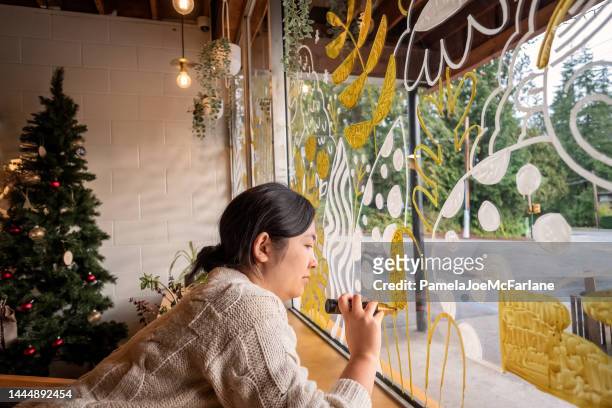 serious eurasian woman painting winter holiday scene on window - store window stock pictures, royalty-free photos & images