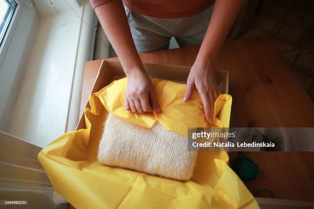 Women's hands pack knitted sweater scarf in yellow paper and cardboard box. Top view.