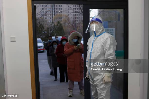 An epidemic control worker wears a protective suit to prevent the spread of COVID-19 as people wait in line for a nucleic acid test on November 27,...