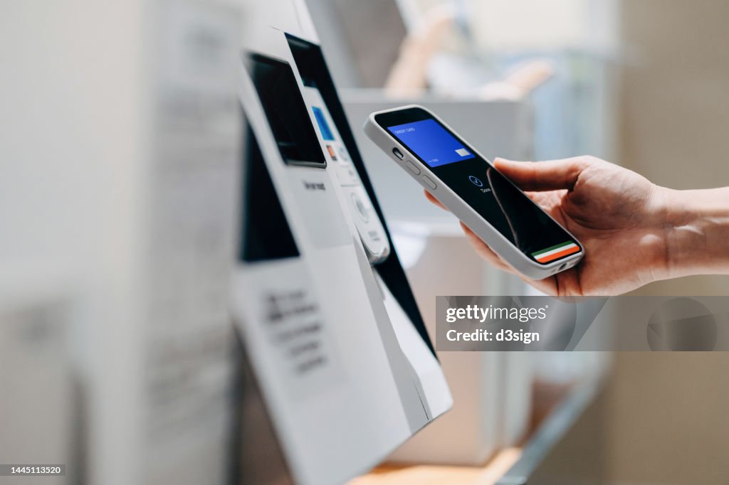 Close up of a woman's hand making mobile payment with her smartphone in a shop, scan and pay a bill on a card machine making a quick and easy contactless payment at self checkout counter. NFC technology, tap and go concept