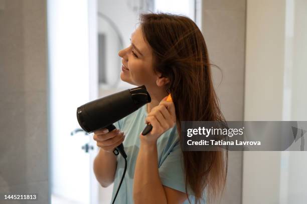 young woman dries her long brown hair with a hair dryer looking to the mirror. attractive female profile view. self care, body care, wellness concept - hair dryer stock pictures, royalty-free photos & images