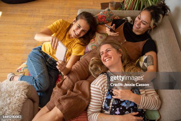 three young women lie together on the sofa looking at their phones - new zealand yellow stock pictures, royalty-free photos & images