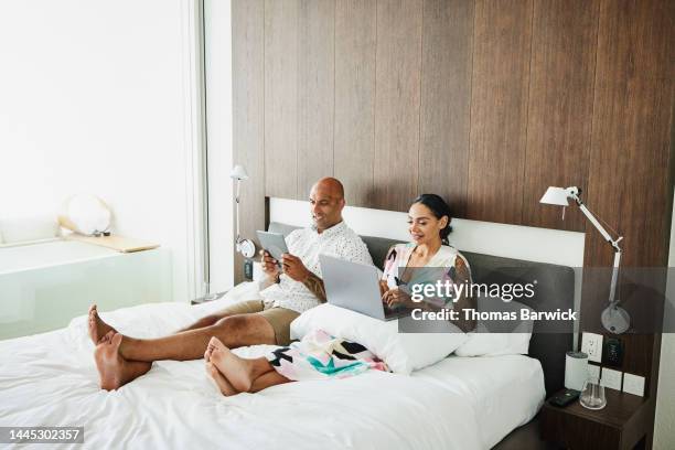 wide shot of couple working on laptop and tablet in bed in hotel room - man in suite holding tablet stock-fotos und bilder