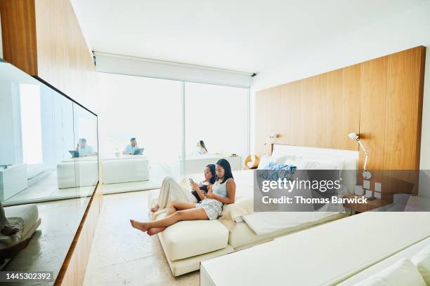 wide shot of sisters looking at smart phones while relaxing in hotel suite - asian man suite stock-fotos und bilder