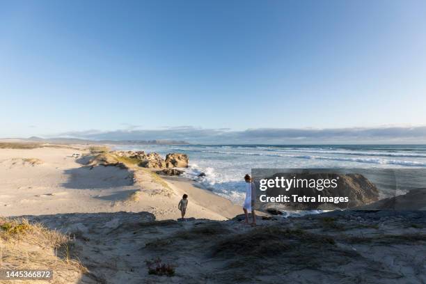 south africa, hermanus, teenage girl (16-17) with younger brother (8-9) climbing on rock on beach - hermanus stock-fotos und bilder
