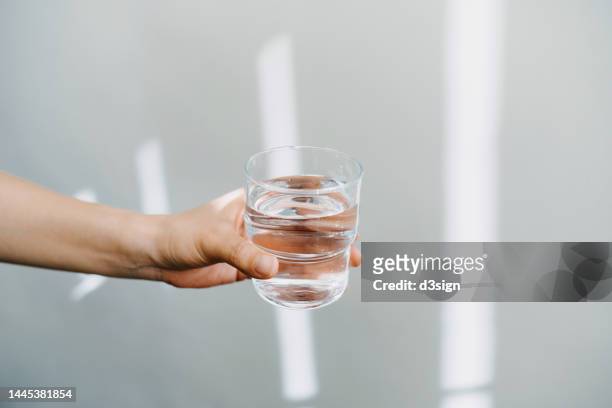 close up of a female hand holding a glass of water against white background with sunlight. healthy lifestyle and stay hydrated - hand glasses stockfoto's en -beelden