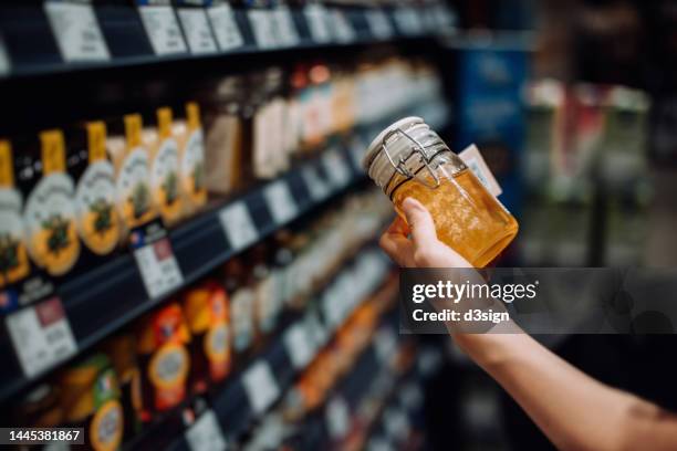 cropped shot of young woman grocery shopping in supermarket, picking up a bottle of organic honey from the product aisle and reading the nutrition label on the bottle. routine shopping. healthy eating lifestyle - honey product shot stock pictures, royalty-free photos & images