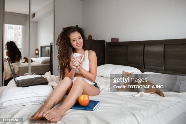 jeune femme métisse buvant du café et travaillant sur des genoux dans sa chambre - cats on the bed photos et images de collection