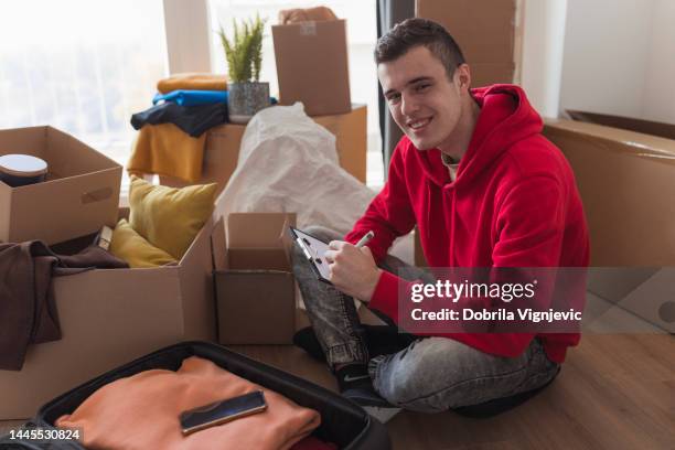 young man sitting among boxes with his belongings, smiling and making to do list - student loan stockfoto's en -beelden