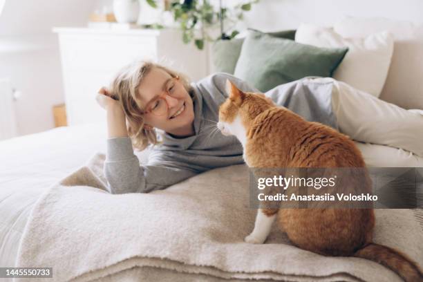 young woman plays with her ginger domestic cat on a bed. cozy relax time - bedclothes stockfoto's en -beelden