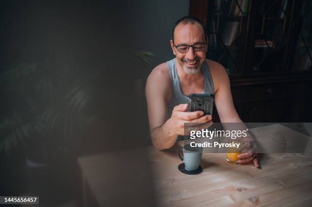 mature man sitting in the kitchen and using smartphone - vruchtensap stockfoto's en -beelden