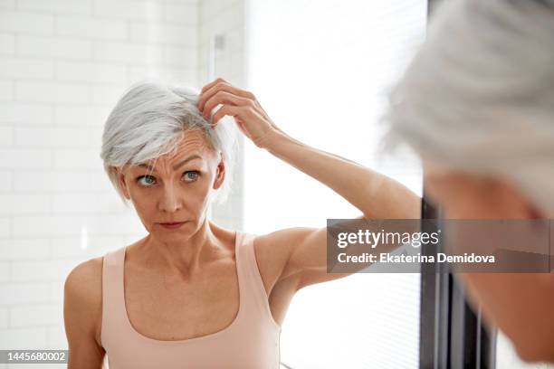 adult woman looking at her gray hair in the mirror - haarausfall stock-fotos und bilder