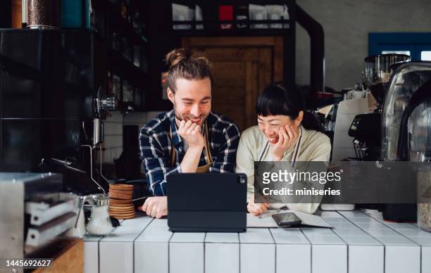 two smiling colleagues using a digital tablet while working together in a coffee shop - friendly small business talking stock pictures, royalty-free photos & images