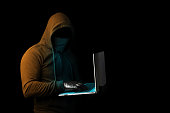 A hooded cyber criminal holding a laptop and scamming online. Cybercrime and phishing alert on the net