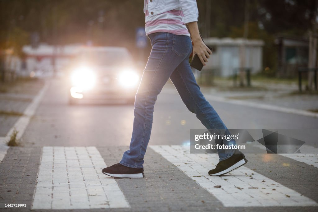 Side view of unrecognizable young woman walking across the crosswalk with car headlights in the background