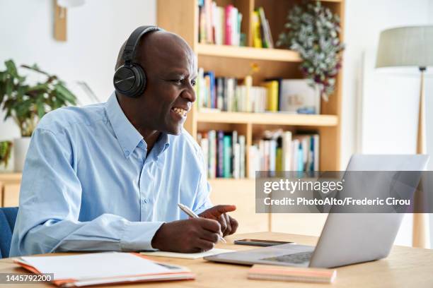 middle-aged african-american man wearing headphones and taking notes while attending remote conference call while working at home with laptop computer - people associated with politics & government stock pictures, royalty-free photos & images