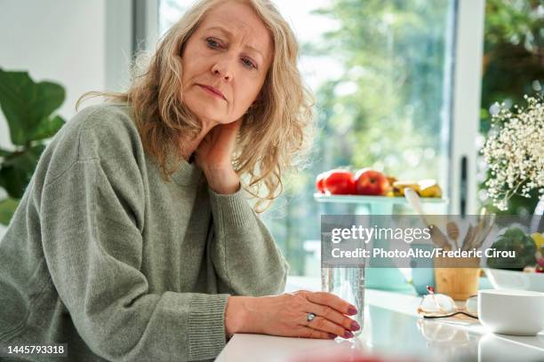 thoughtful senior woman with hand behind neck sitting in dining room - ongerust stockfoto's en -beelden