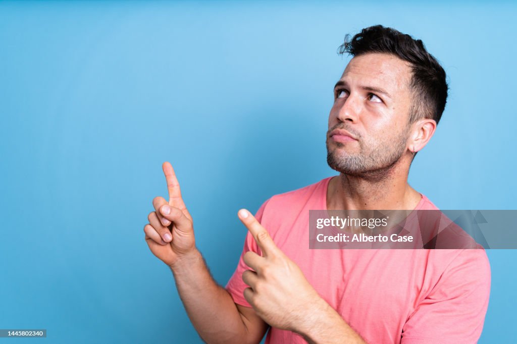 Man with a doubtful face looking at an empty space and pointing with his hands on a blue background. Perfect for copy space