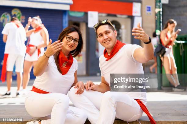 man and woman sitting on the sidewalk smiling at the camera with glasses on their heads and waving, front view in traditional san fermines costume - pamplona fotografías e imágenes de stock