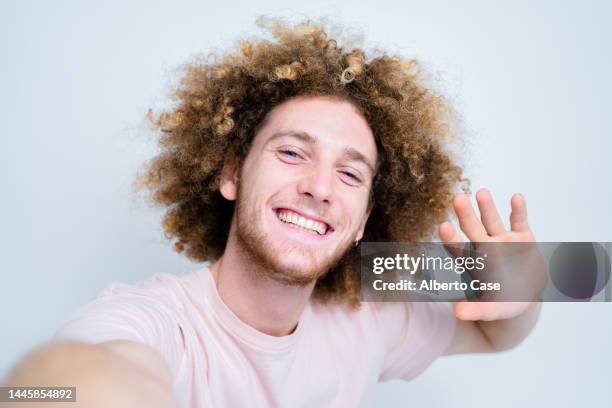 happy curly-haired man taking a selfie with his phone and waving on a white background - homme enthousiasme sourire fond blanc photos et images de collection