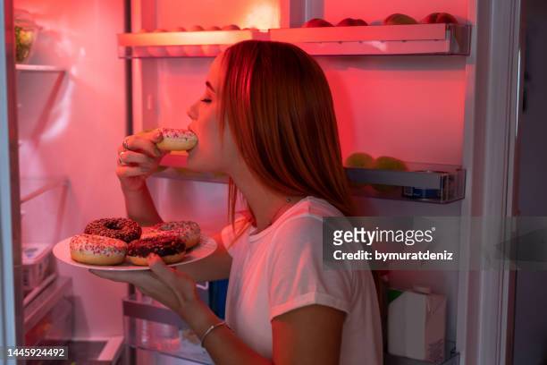 donna che mangia ciambelle malsane davanti al frigorifero aperto - eating donuts foto e immagini stock