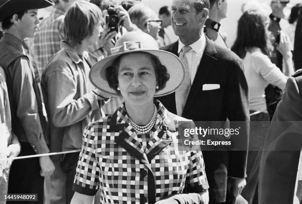 British Royal Queen Elizabeth II , wearing a checked jacket with a pearl necklace, and a hat with a buckle on the front, with her husband, Prince...