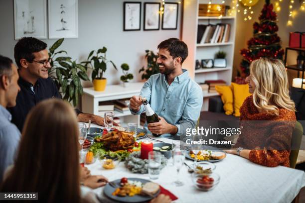 group of diverse friends enjoying christmas dinner at home, young man opening champagne bottle - opening night dinner stockfoto's en -beelden