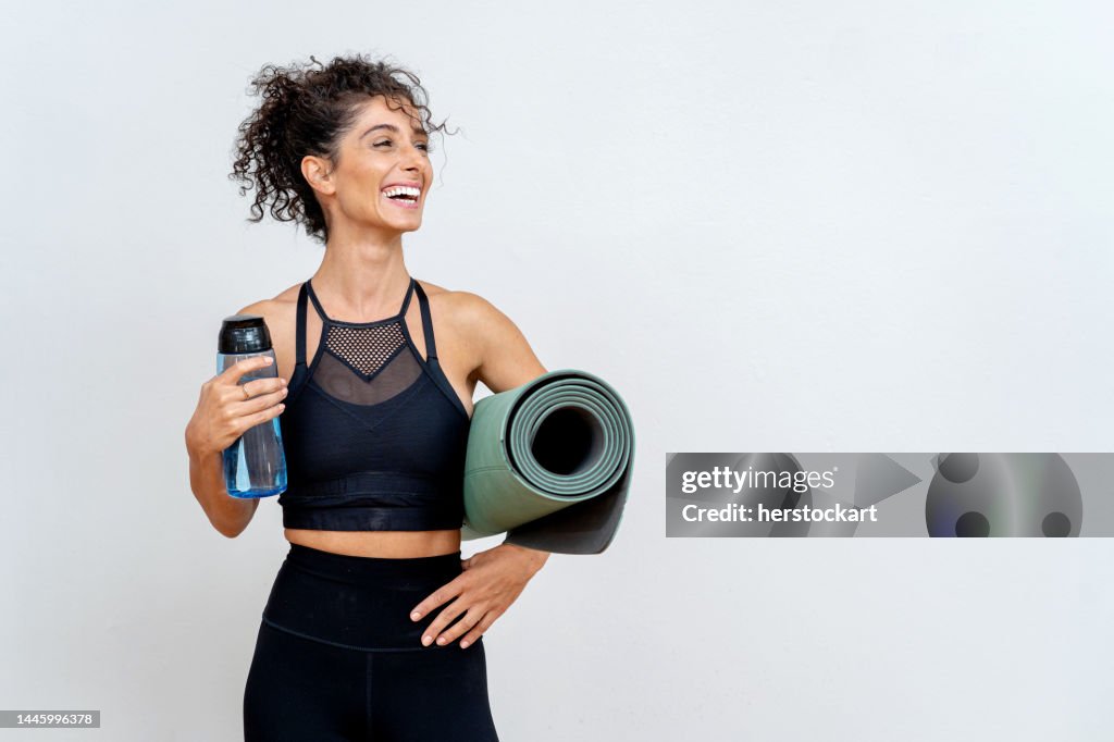 Femme souriante devant un mur blanc avec tapis et bouteille d’eau