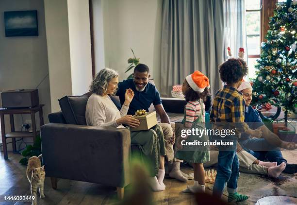 gift, christmas and big family happy to celebrate, holiday diversity and giving in the living room of a house. festive, celebrating and senior woman opening a present for xmas in an interracial home - grandmas living room stockfoto's en -beelden