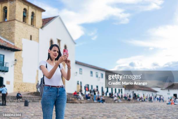 latina-frau ist in der mitte des platzes in villa de leyva mit ihrem handy und macht einen videoanruf mit ihrer familie - villa de leyva stock-fotos und bilder