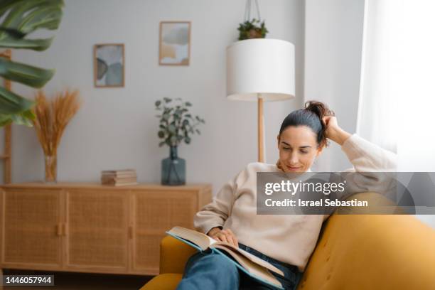 view of a woman, dressed in casual clothes, sitting in a comfortable and carefree position on a yellow sofa in the bright living room of her house, smiling while reading a book. - contemporary literature stock pictures, royalty-free photos & images