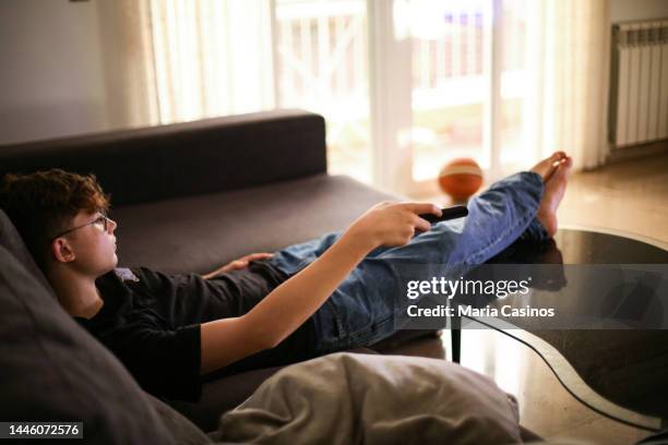 teen boy lying  down  on the sofa holding the tv remonte - teen boy barefoot stock pictures, royalty-free photos & images