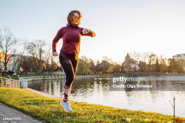 checking heartbeat and pressure during jogging. mature red haired female running near lake in green park - watch time stock pictures, royalty-free photos & images