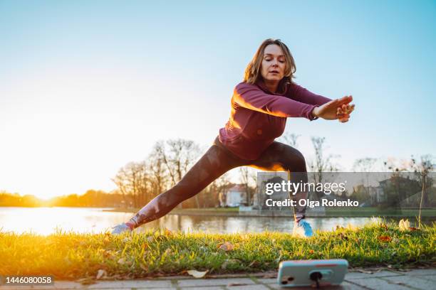 learning yoga and fitness positions from the internet on the phone. athlete woman making squats for glutes and leg muscles in green park - woman stretching sunset stock-fotos und bilder