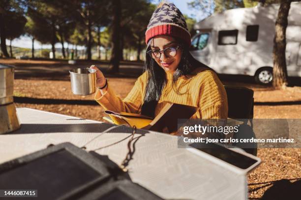 hispanic woman reading a book at a campsite while charging her smartphone with a solar panel - drinking coffee - - alternativer lebensstil stock-fotos und bilder