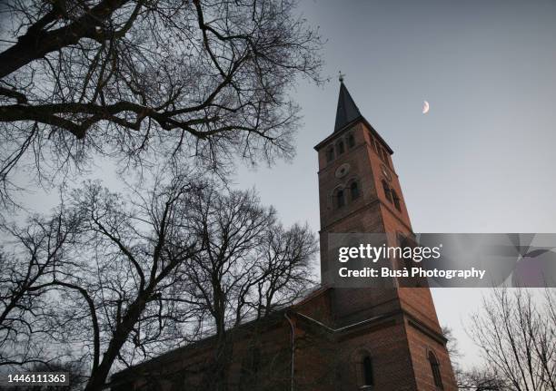 bell tower of rural gothic church with bare winter trees and moon at night - brick cathedral stock pictures, royalty-free photos & images