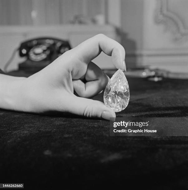 Close-up of a woman's hand holding a large pear-shaped diamond between the thumb and forefinger, a telephone visible in the background, United...