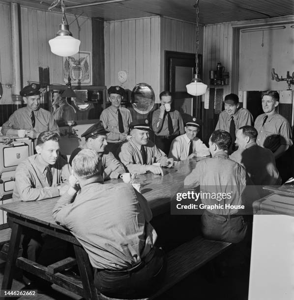 Group of firefighters in dress uniform in the canteen, some standing while others sit at a table, at an fire station, United States, circa 1945.