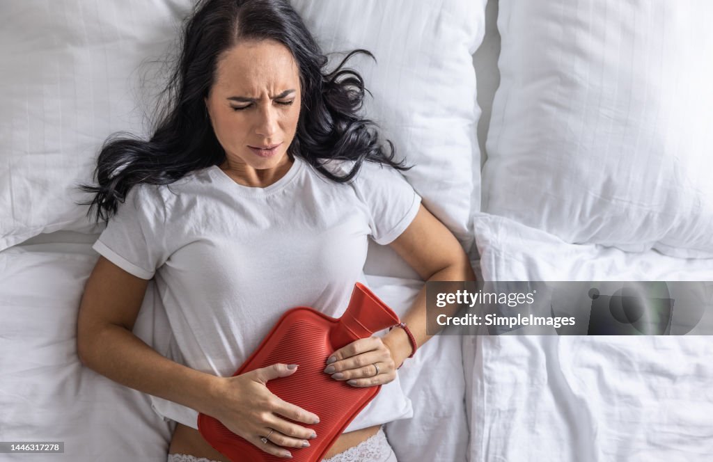 A young woman suffers from menstrual pains, lies on the bed and warms her stomach with a rubber heating pad.
