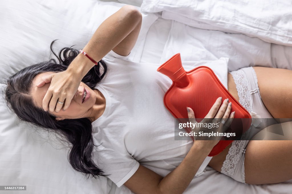 A young woman suffers from menstrual pains, lies on the bed and warms her stomach with a rubber heating pad.