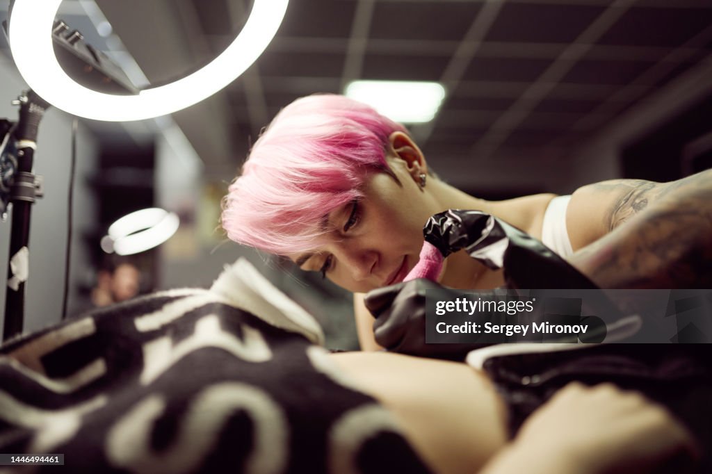 Young woman with pink hair tattooing client in own studio