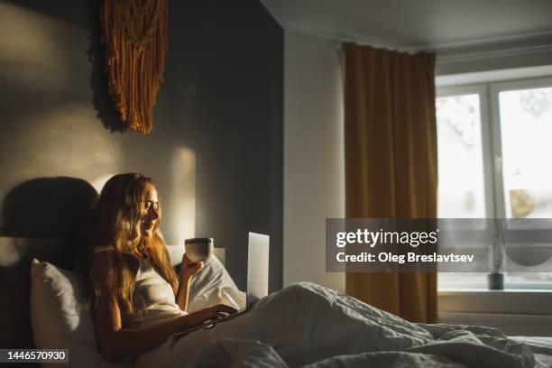 woman leisure in bed with cup of coffee and laptop. beautiful morning light from window - women drinking coffee photos et images de collection