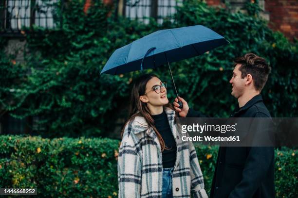 young man giving umbrella to girlfriend in front of plants - sharing umbrella stock pictures, royalty-free photos & images