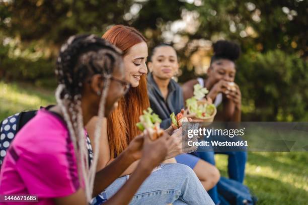 smiling young woman with friends eating snack sitting in park - university student picnic stock pictures, royalty-free photos & images