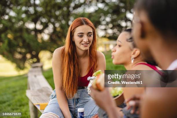 young woman looking at friend sitting with food in park - university student picnic stock pictures, royalty-free photos & images