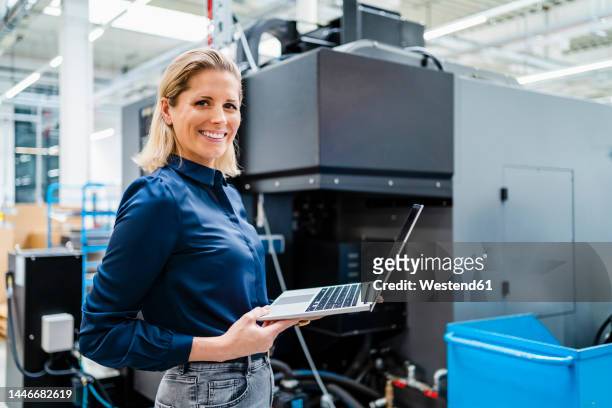 smiling businesswoman with laptop standing by machinery in factory - blue blouse stock pictures, royalty-free photos & images